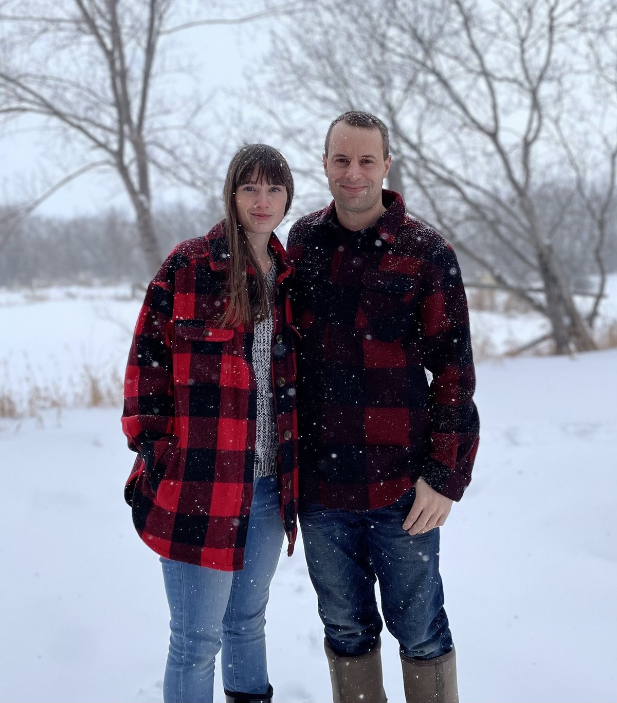 Jed and Lucia Janzen standing together in falling snow on their farm near Rosthern, Saskatchewan, both wearing red buffalo-plaid jackets